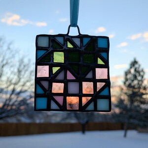 以下が含まれることがあります： 幾何学模様のステンドグラスのサンキャッチャー。青、緑、ピンク、透明ガラスの正方形と三角形のデザインで、黒いハンダで縁取られています。吊り下げ用の水色のリボンが付いています。
