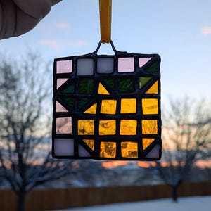 May include: A square stained glass ornament with a geometric design. The ornament features a mix of colors, including pink, white, green, and yellow. It is suspended by a yellow ribbon. The background shows a winter scene with a sunset.