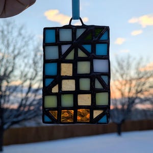 以下が含まれることがあります： 幾何学模様の長方形ステンドグラスサンキャッチャー。青、金、緑、白の色合いの正方形と三角形が特徴で、黒い線で縁取られています。吊り下げ用の水色のリボンが付いています。