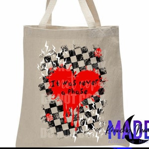 May include: A white tote bag with a black and white checkered pattern and a red heart with the text "It was never a phase" in black. The heart is dripping red paint and there are white lightning bolts around the checkered pattern. The bag has a black and white logo that says "Krunchy Mama Made" with the words "Adaptable + More" underneath.