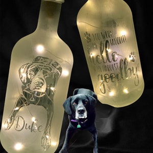 May include: Two frosted glass bottles with lights inside. One bottle has a silhouette of a dog with the name "Duke" etched on it. The other bottle has the text "You were my favourite hello and my hardest goodbye" etched on it. A black dog is sitting in front of the bottles.