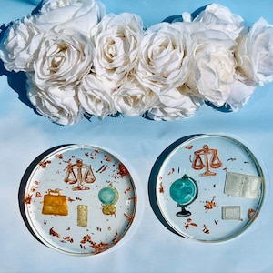 May include: Two round coasters with a gold and white design. One coaster features a scale of justice, a globe, and a book. The other coaster features a scale of justice, a globe, and a briefcase.