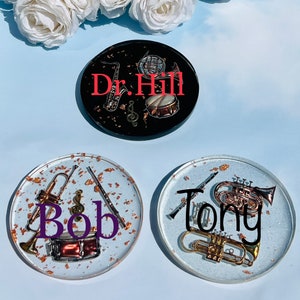 May include: Three round coasters with a white background and gold glitter. Each coaster features a different musical instrument and a name. The first coaster has a saxophone, a drum, and the name "Dr. Hill". The second coaster has a trumpet, a flute, and the name "Bob". The third coaster has a french horn, a trumpet, and the name "Tony".