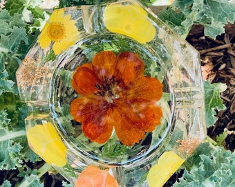May include: A clear glass ashtray with a pressed flower design. The ashtray features a large orange flower in the center, surrounded by smaller yellow and orange flowers. The ashtray is octagonal in shape.