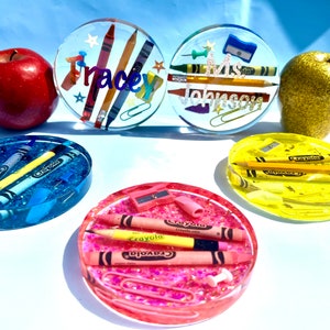 May include: Five round resin coasters with different colors and school supplies embedded inside. The coasters are blue, yellow, pink, clear with the name "Tracey" and clear with the name "Mrs. Johnson".