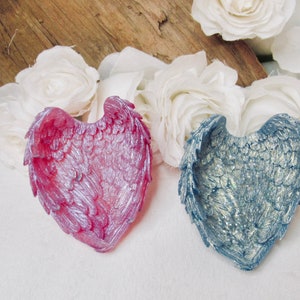 May include: Two heart-shaped decorative items with a wing design. One is a pink and silver color, the other is a blue and silver color.