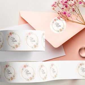 Custom sticker labels displayed on a roll beside a blush envelope, floral accents, and wedding rings on a neutral surface, showing round stickers used for stationery sealing and event packaging.