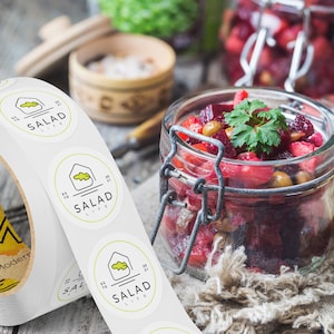 Custom sticker labels displayed on a roll beside a glass jar filled with fresh salad ingredients on a rustic tabletop, showing round food labels applied to packaging in a kitchen setting.