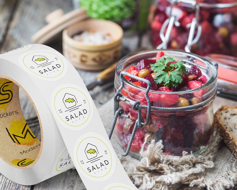 Custom sticker labels displayed on a roll beside a glass jar filled with fresh salad ingredients on a rustic tabletop, showing round food labels applied to packaging in a kitchen setting.