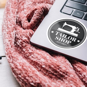May include: A black and white sticker with a sewing machine graphic and the text "Tailor Shop - Est. 1987" on a silver laptop.