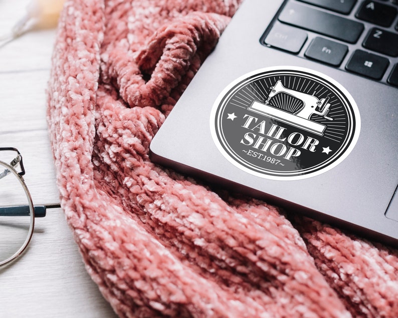 May include: A black and white sticker with a sewing machine graphic and the text "Tailor Shop - Est. 1987" on a silver laptop.