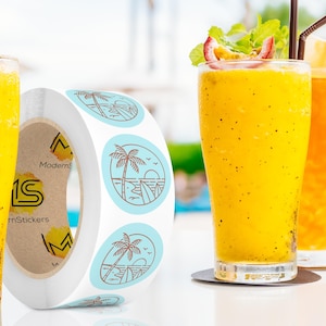Custom sticker labels displayed on a roll beside tall glasses of yellow beverages with garnishes on a light tabletop, showing round logo stickers in roll format.