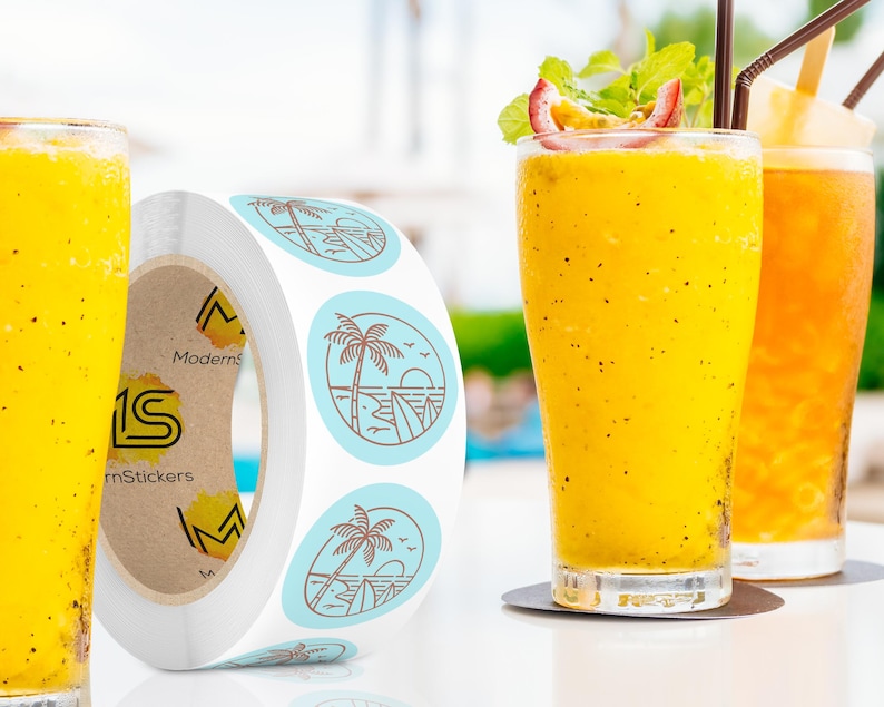 Custom sticker labels displayed on a roll beside tall glasses of yellow beverages with garnishes on a light tabletop, showing round logo stickers in roll format.