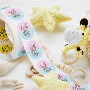 Custom sticker labels displayed on a roll beside soft baby toys and crocheted accessories on a light fabric surface, showing round pastel logo stickers in roll format.