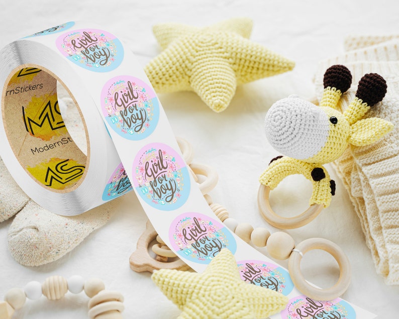 Custom sticker labels displayed on a roll beside soft baby toys and crocheted accessories on a light fabric surface, showing round pastel logo stickers in roll format.
