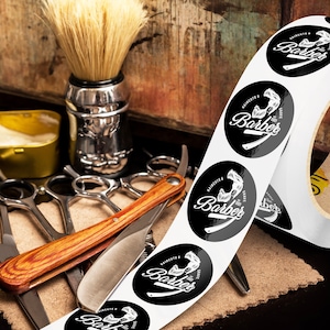 Custom sticker labels displayed on a roll beside barber tools including scissors, shaving brush, razor, and grooming accessories on a rustic work surface, showing round logo stickers in roll format.