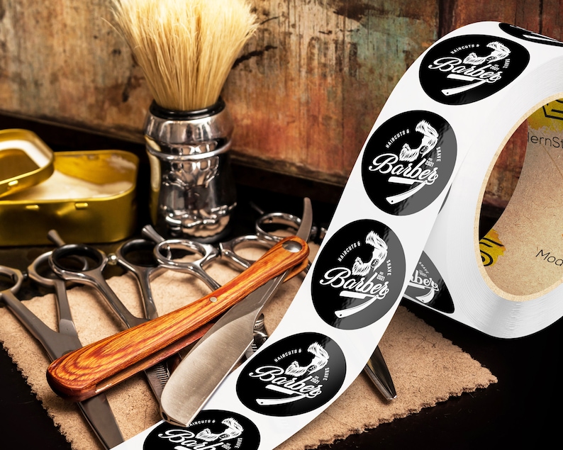 Custom sticker labels displayed on a roll beside barber tools including scissors, shaving brush, razor, and grooming accessories on a rustic work surface, showing round logo stickers in roll format.