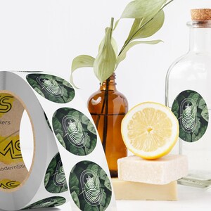 Custom sticker labels displayed on a roll beside glass bottles, a sliced lemon, and natural decor on a white surface, showing round logo stickers in roll format.