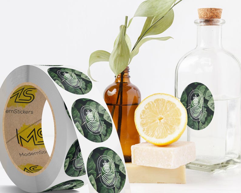 Custom sticker labels displayed on a roll beside glass bottles, a sliced lemon, and natural decor on a white surface, showing round logo stickers in roll format.
