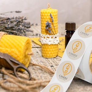 Custom sticker labels displayed on a roll beside beeswax candles, scissors, dried florals, and twine on a neutral tabletop, showing round logo stickers in roll format.