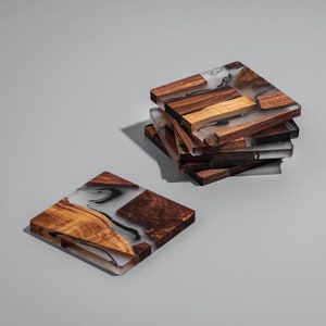 May include: A stack of square coasters made of wood and clear resin. The wood pieces are various shades of brown and tan, while the resin has a marbled, smoky appearance. One coaster is separate from the stack, lying flat.