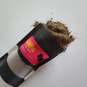 May include: A black pouch with a pink and yellow label that says "NO-NATURE CHEBE powder" and a picture of a woman with curly hair. The pouch is open and brown powder is spilling out.