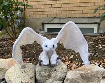 May include: A white crochet dragon with large, outstretched wings. The dragon has blue eyes and gray accents on its feet. It is sitting on a rock, with a brick wall and greenery in the background. A handmade, whimsical item.