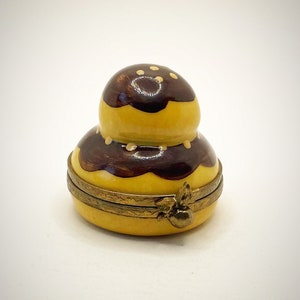 Pode incluir: Uma pequena caixa de porcelana redonda, amarela e marrom, com uma tampa articulada. A caixa é decorada com um desenho de um pastel com cobertura de chocolate e granulado branco.