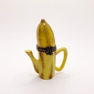 May include: A yellow ceramic teapot shaped like a banana with a brown metal lid and handle. The teapot has a brown and yellow striped pattern.