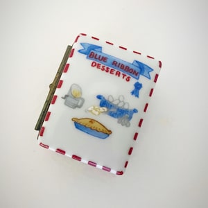 Puede incluir: Una pequeña caja rectangular con una superficie blanca y un borde de puntos rojos y blancos. La caja presenta una pancarta azul con las palabras "BLUE RIBBON DESSERTS" e ilustraciones de postres.