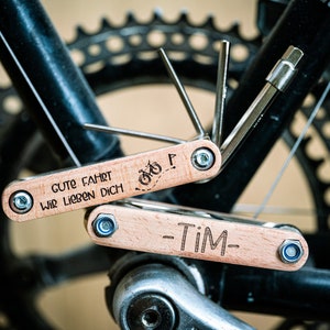 Könnte beinhalten: Zwei Holz-Multitools mit einem Fahrrad-Symbol und dem Text "Gute Fahrt Wir Lieben Dich" auf einem Werkzeug und "Tim" auf dem anderen.