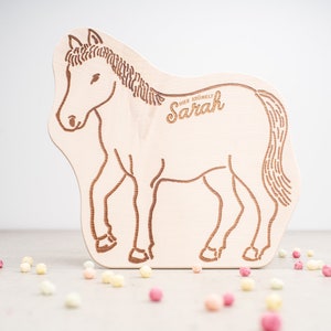 Könnte beinhalten: Holzpferd-förmige Plakette mit einem geätzten Design. Das Pferd ist hellbraun, und der Name "Sarah" ist kursiv geätzt. Die Plakette befindet sich auf einer hellgrauen Oberfläche mit bunten Süßigkeiten.