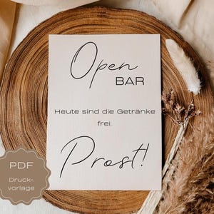 Könnte beinhalten: Ein beiges Schild mit schwarzer Schrift, das "Open BAR" und "Prost!" in einer kursiven Schriftart zeigt. Das Schild befindet sich auf einer Holzscheibe mit getrockneten Pflanzen und einer Feder. Der Text "Heute sind die Getränke frei" steht ebenfalls auf dem Schild.