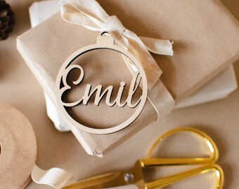 Könnte beinhalten: Ein runder Weihnachtsbaumschmuck aus Holz mit dem Namen "Emil", der in einer Schreibschrift ausgeschnitten ist. Der Schmuck liegt auf einem braunen Geschenkpaket mit einem weißen Band.