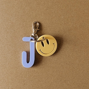 Könnte beinhalten: Ein Schlüsselanhänger mit einem silbernen Verschluss, einem hellblauen Buchstaben "J" und einem gelben Smiley-Gesicht. Das Smiley-Gesicht hat schwarze Augen und einen gebogenen Mund. Der Schlüsselanhänger befindet sich auf braunem Hintergrund.