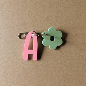 Könnte beinhalten: Ein rosa Schlüsselanhänger mit dem Buchstaben "A" und ein hellgrüner blütenförmiger Anhänger sind durch einen silberfarbenen Ring verbunden. Der Schlüsselanhänger hat einen silbernen Verschluss. Der Hintergrund ist einfarbig beige.