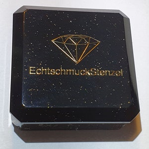 May include: Black jewellery box with a gold diamond graphic and the text "EchtschmuckStenzel". The box has a faceted design and a speckled finish, suggesting a luxurious aesthetic. The box is likely for storing jewellery.