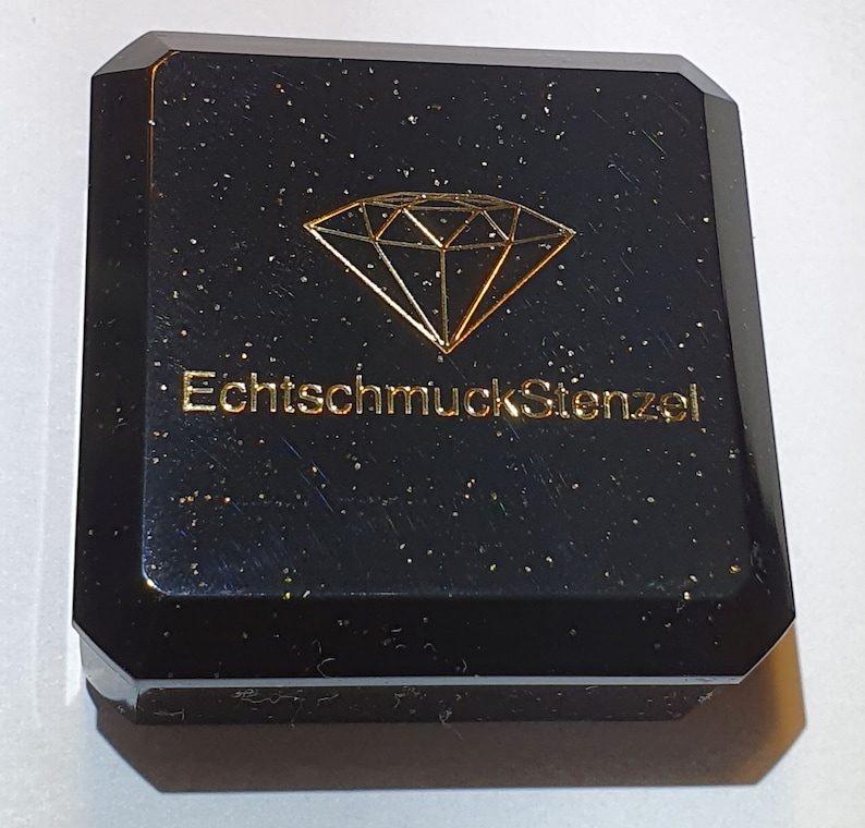 May include: Black jewellery box with a gold diamond graphic and the text "EchtschmuckStenzel". The box has a faceted design and a speckled finish, suggesting a luxurious aesthetic. The box is likely for storing jewellery.