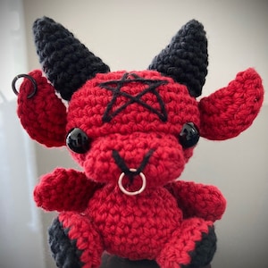 Puede incluir: Un juguete de peluche de diablo rojo tejido a crochet con cuernos negros, un pentagrama en la frente y un anillo plateado en la nariz.