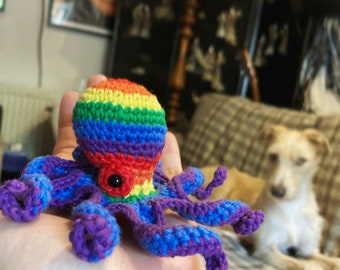 Puede incluir: Un juguete de pulpo de crochet con rayas de colores arcoíris y un solo ojo negro. El pulpo está sentado en la mano de una persona.