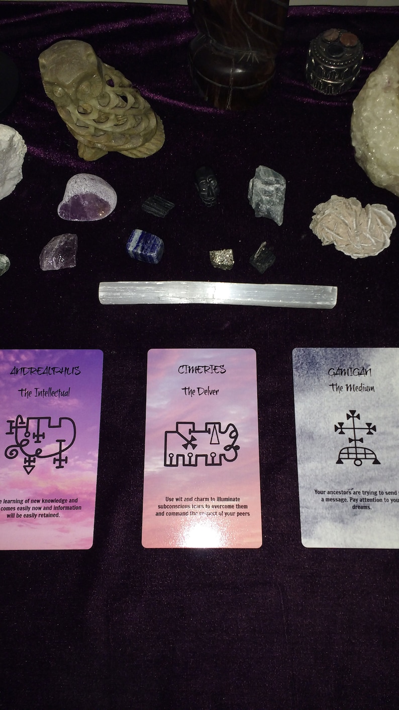 Puede incluir: Tres cartas de or&aacute;culo rectangulares con los nombres Andrealphus, Cimeries y Gamigin con sellos y texto en negro, dispuestas bajo una colecci&oacute;n de varios cristales en bruto, piedras y una figura de b&uacute;ho de piedra sobre tela morada oscura.