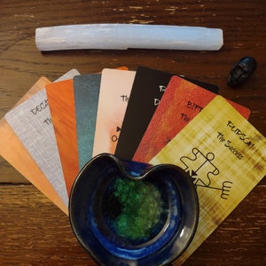 Puede incluir: Un abanico de cartas de tarot de varios colores con texto que incluye Purson the Success, un cuenco de cer&aacute;mica azul en forma de coraz&oacute;n lleno de cristales verdes, una varita de selenita blanca larga y una peque&ntilde;a figura de calavera negra.