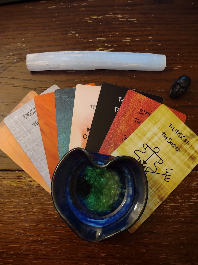 Puede incluir: Un abanico de cartas de tarot de varios colores con texto que incluye Purson the Success, un cuenco de cer&aacute;mica azul en forma de coraz&oacute;n lleno de cristales verdes, una varita de selenita blanca larga y una peque&ntilde;a figura de calavera negra.