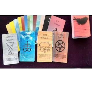 Puede incluir: Una baraja de cartas or&aacute;culo rectangulares con diversos colores y s&iacute;mbolos. Cuatro cartas est&aacute;n desplegadas en primer plano, mostrando los nombres Lucifer, Leviathan, Belial y Satan, junto con sus respectivos sellos y textos descriptivos.