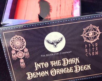 May include: A black box with a gold and white design. The box has the text "Into the Dark Demon Oracle Deck" and "The Silver Owl Company" on the front. The box is open and shows a red interior.