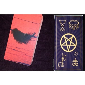 Puede incluir: Una baraja de cartas con reverso rojo que muestra una pluma negra y una caja morada oscura con un pentagrama dorado, s&iacute;mbolos ocultos y una luna creciente. Las cartas tienen esquinas redondeadas y un acabado liso junto a la caja.