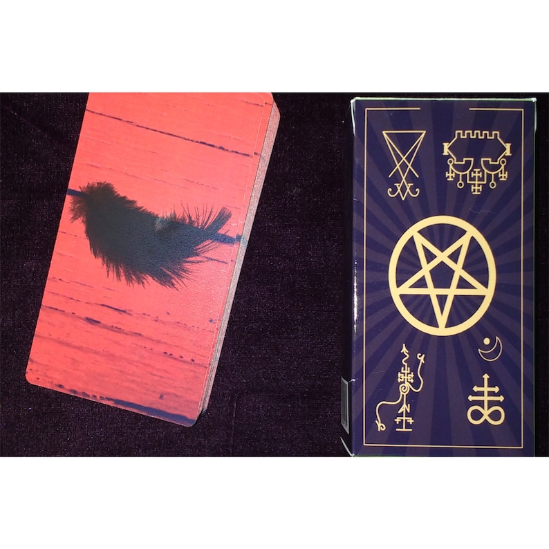 Puede incluir: Una baraja de cartas con reverso rojo que muestra una pluma negra y una caja morada oscura con un pentagrama dorado, s&iacute;mbolos ocultos y una luna creciente. Las cartas tienen esquinas redondeadas y un acabado liso junto a la caja.