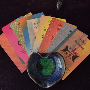 Puede incluir: Un abanico de cartas de or&aacute;culo coloridas con varios dise&ntilde;os y texto, incluyendo Hekate the Doorway, dispuestas detr&aacute;s de un plato de cer&aacute;mica en forma de coraz&oacute;n con un exterior azul profundo y un interior cristalino verde vibrante.