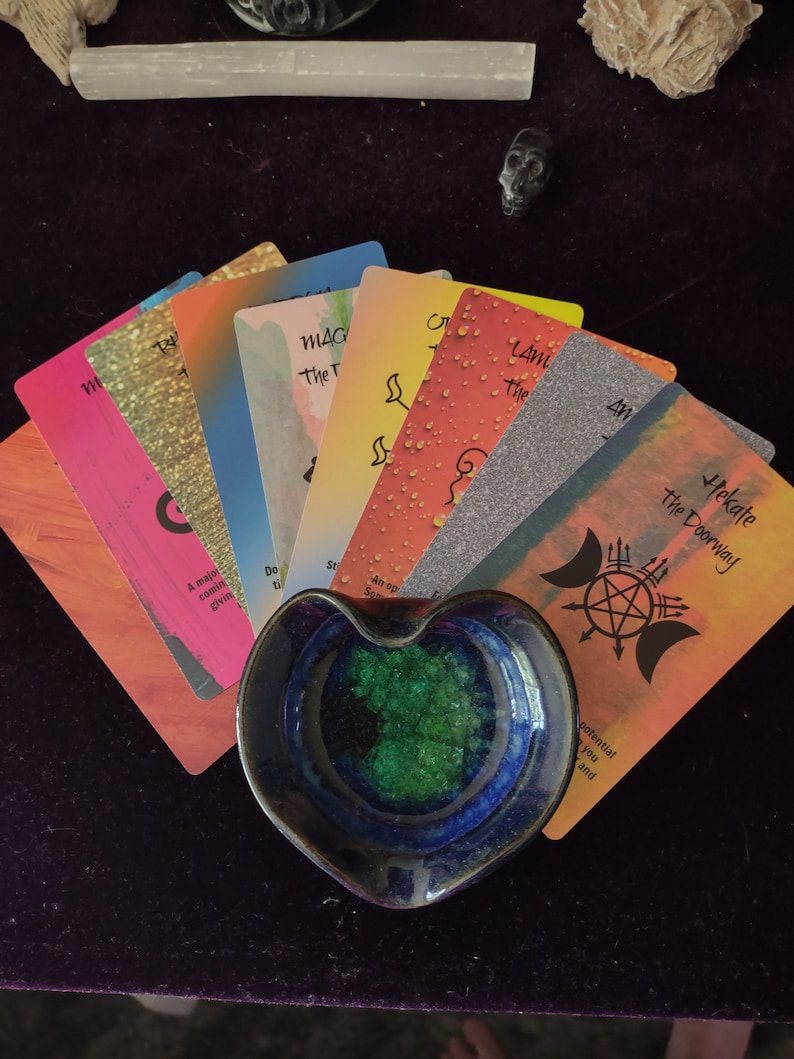 Puede incluir: Un abanico de cartas de or&aacute;culo coloridas con varios dise&ntilde;os y texto, incluyendo Hekate the Doorway, dispuestas detr&aacute;s de un plato de cer&aacute;mica en forma de coraz&oacute;n con un exterior azul profundo y un interior cristalino verde vibrante.