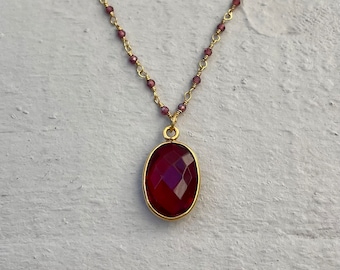 May include: A gold-colored necklace with a large, oval, faceted, deep red pendant. The pendant is set in a gold bezel. The necklace chain features small, round, red beads. The necklace is displayed on a white, textured surface.